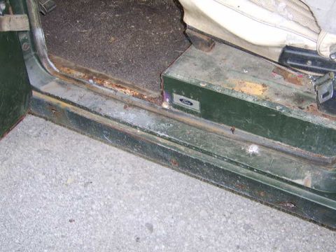 1976 Ford Bronco - The interior of a vehicle shows significant rust around the door sill and a visible Ford emblem on a green panel.