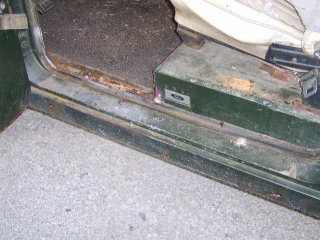 Close-up of a vehicle's rusty door sill and floor mat, with a Ford emblem visible on a green metal panel