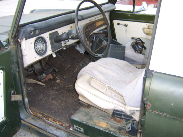 Green vehicle interior showing dashboard, steering wheel, pedals, and worn seat
