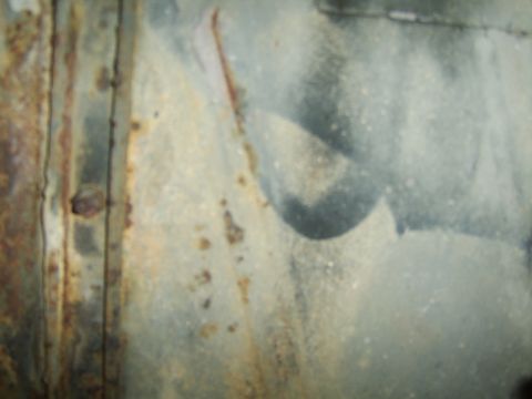 1976 Ford Bronco - This image shows a close-up of a heavily rusted metal panel with some gray paint splatters.