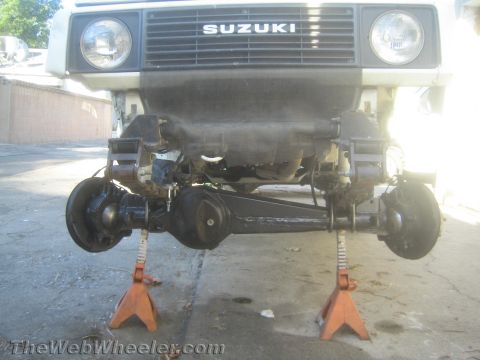 1987 Suzuki Samurai - Front view of a Suzuki 4x4 raised on two orange jack stands, showing the front live axle, differential, leaf springs, and brake drums with…