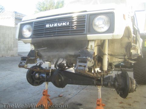1987 Suzuki Samurai - The front axle and leaf spring suspension of a white 1987 Suzuki Samurai are visible, supported by jack stands.