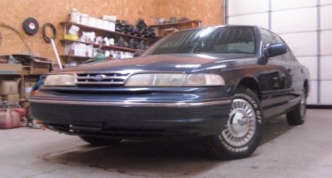 1997 Ford Crown Victoria - A dark blue sedan is parked inside a garage with shelves of supplies in the background.