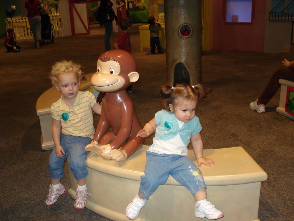 Two children sit with a Curious George statue in a museum exhibit