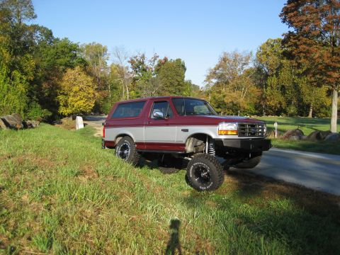 1996 Ford Bronco - A lifted Ford Bronco sits on the side of a road with trees in the background.