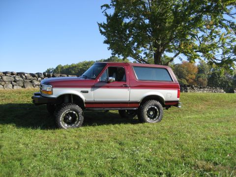 1996 Ford Bronco - This two-tone 1996 Ford Bronco is seen from the side, parked in a grassy field with a stone wall and trees in the background.