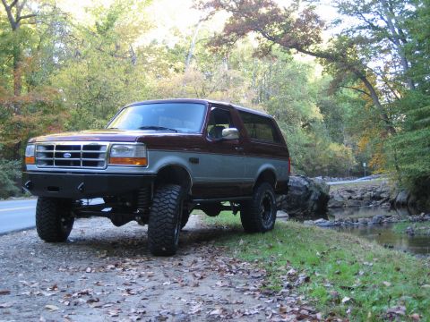 1996 Ford Bronco - 1996 Ford Bronco parked off-road during autumn with trees and a stream visible.