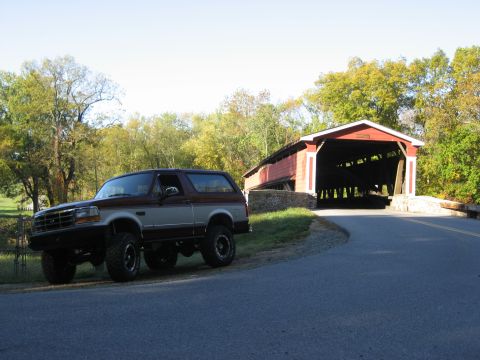 1996 Ford Bronco - A two-tone Ford Bronco sits beside a historic covered bridge on a sunny day.