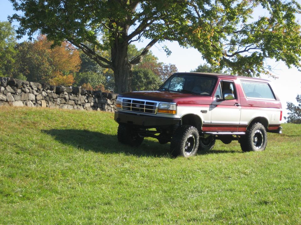 Maroon and white Ford Bronco parked on a grassy hill with a stone wall behind it
