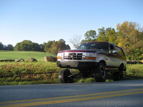 1996 Ford Bronco - This lifted 1996 Ford Bronco sits on a grassy shoulder next to a road, with trees and a field in the background.