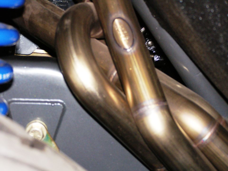 Close-up of exhaust header tubes with weld seams and an oval stamp in an engine bay