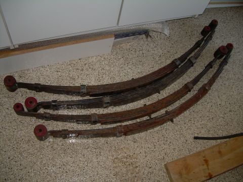 1986 Ford Bronco - Four rusty leaf springs with red bushings are laid out on a speckled floor.