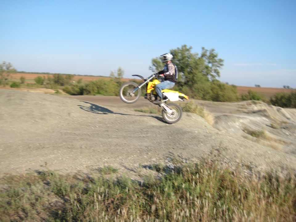 Motorcyclist performing a wheelie on a dirt track with dry grass and trees in the background