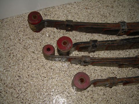 1986 Ford Bronco - Several leaf springs with red bushings are laid out on a speckled surface. A website logo is visible in the lower right corner.