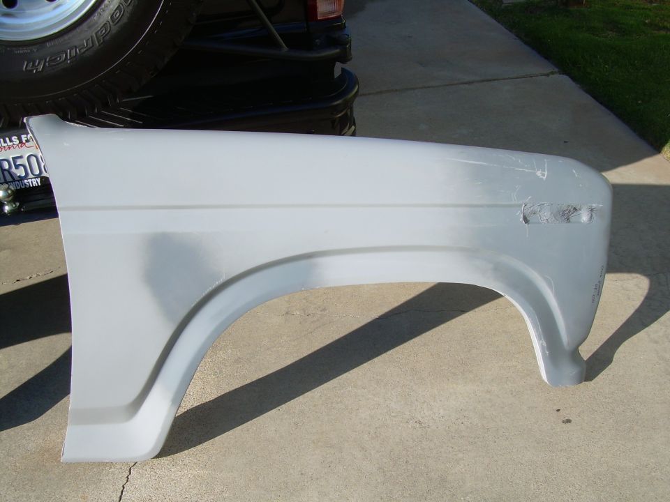 Gray fender part sitting on concrete, with a tire and part of a vehicle in the background. Supermotors.net logo visible
