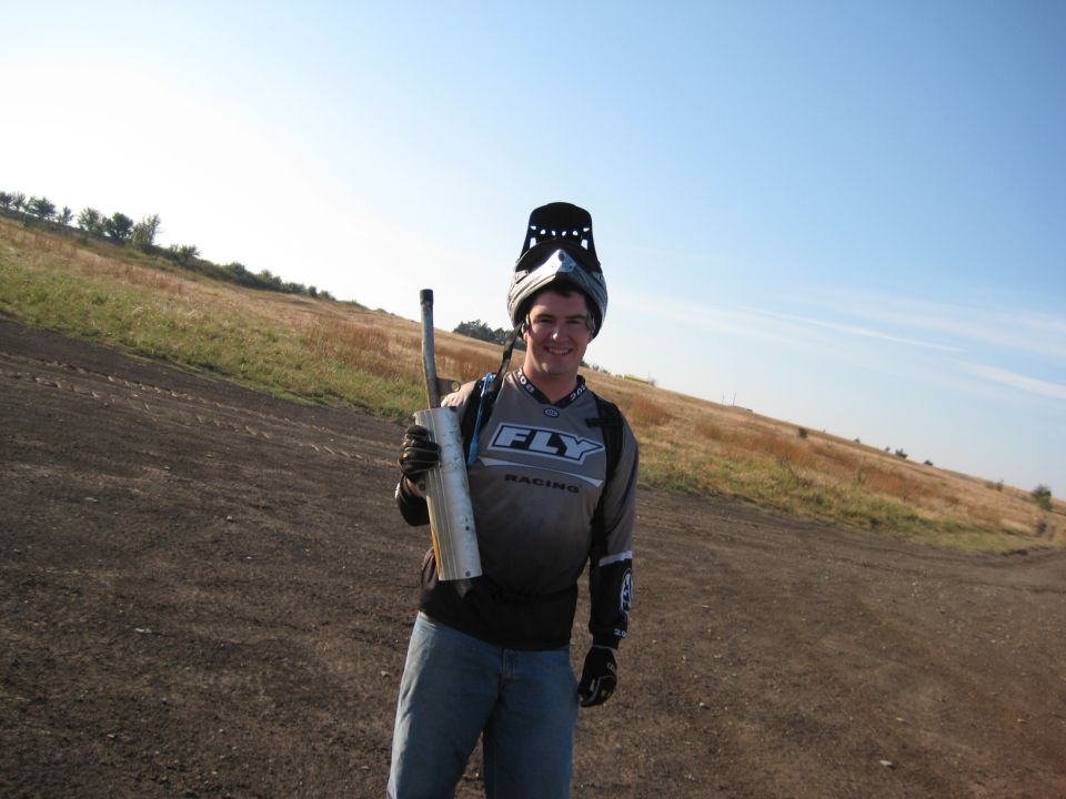 Guy in motocross gear holding a dirt bike exhaust pipe in a dry, grassy field