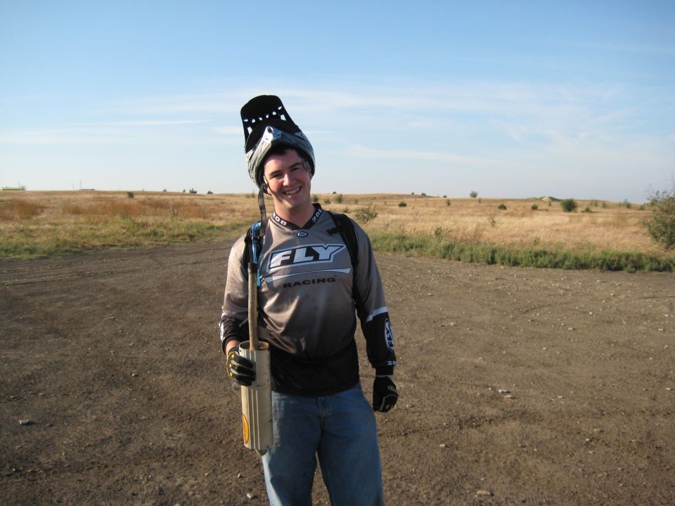 Man in motocross gear holding a pipe, standing in a dry, grassy field under a clear blue sky