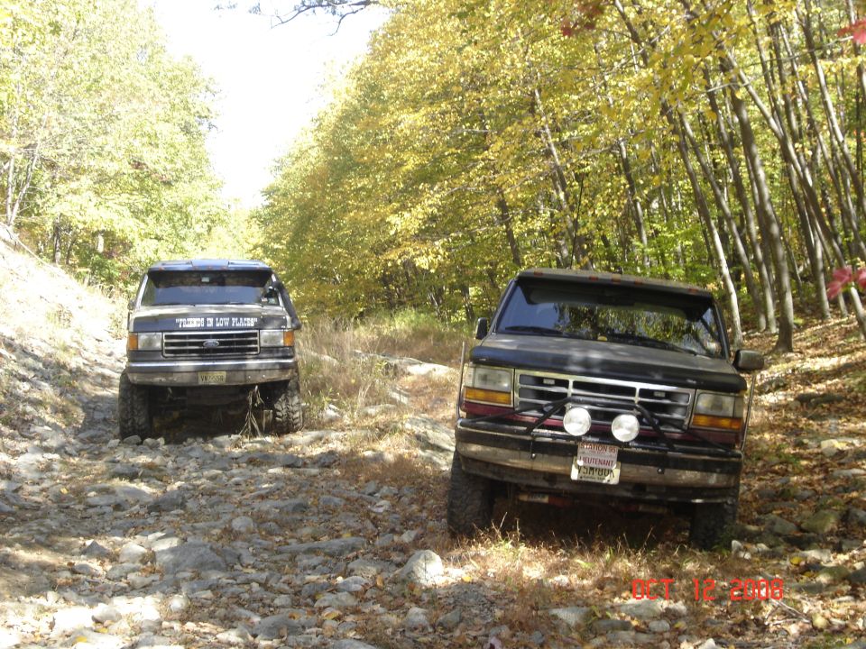 Two dark Ford Broncos with lifted suspensions drive on a rocky, wooded trail in autumn. The front Bronco has a license plate and two fog lights. The rear Bronco has text on its windshield