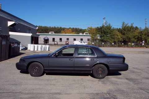 2001 Ford Crown Victoria - A dark sedan is parked in an industrial lot under a clear blue sky. Numbers are written on the rear window.