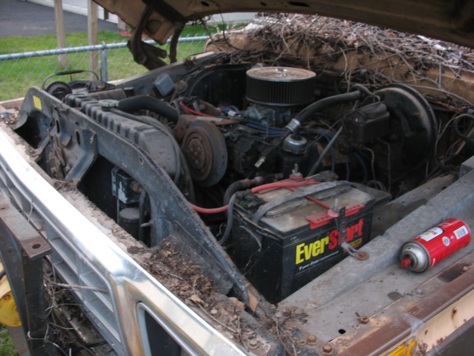 Truck engine bay with a V8 engine, battery, and radiator. The hood is open, and debris covers the engine. A red spray can is on the fender