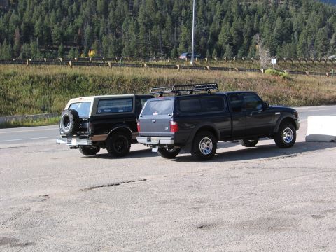 1996 Ford Bronco - Two SUVs are parked side-by-side in a gravel lot with a wooded hillside in the background. The SUV on the left is a black Ford Bronco with…
