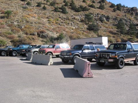 1996 Ford Bronco - A lineup of SUVs and pickup trucks are parked in a lot with a rocky, tree-covered hillside in the background. Several concrete barriers are…