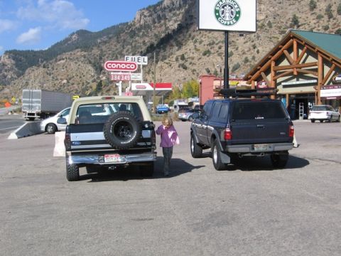 1996 Ford Bronco - The rear view of a tan and black Ford Bronco with a spare tire mounted on the tailgate is parked next to a dark blue pickup truck with a ca…