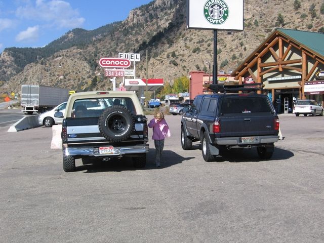 Tan and black Ford Bronco and dark blue pickup truck parked in a lot with mountains and Starbucks in the background.