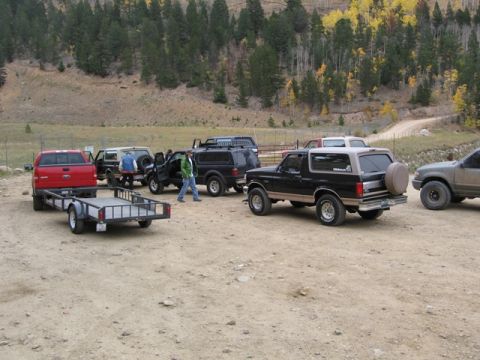 1996 Ford Bronco - A black and tan Ford Bronco is parked on a dirt lot with several other vehicles and a trailer. The Bronco has a spare tire mounted on the r…
