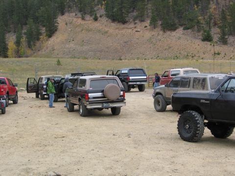 1996 Ford Bronco - Several off-road vehicles are parked on a dirt lot with a wooded hillside in the background. A black Ford Bronco with a spare tire on the b…