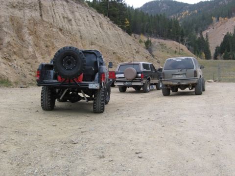 1996 Ford Bronco - The rear of a lifted black pickup truck with a spare tire mounted on the back is parked on a dirt lot with two SUVs behind it. The scene is…