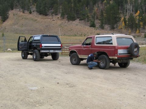 1996 Ford Bronco - A man is working on the rear tire of a red and tan Ford Bronco. Another black Ford Bronco with its driver's side door open is parked to the…