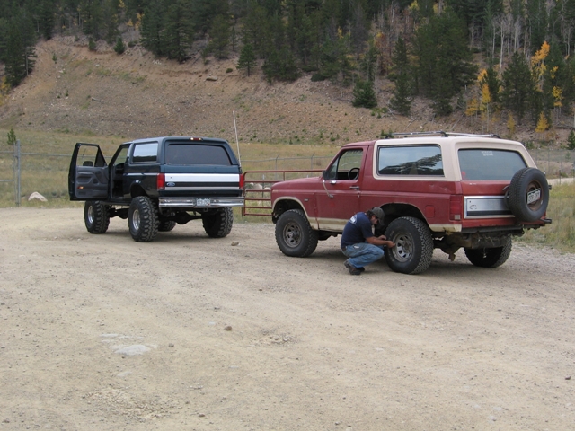Two Ford Broncos parked on a dirt road with a man working on the rear tire of the red Bronco.