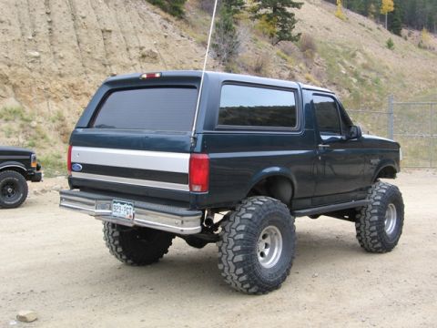 1996 Ford Bronco - The rear driver's side quarter view of a lifted, dark green 1996 Ford Bronco with large off-road tires is shown parked on a dirt road with…