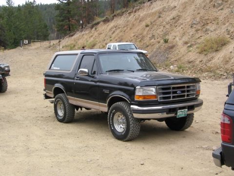 1996 Ford Bronco - A black 1990s Ford Bronco with a tan lower body stripe is parked on a dirt road in a hilly, wooded area. The SUV has aggressive all-terrain…