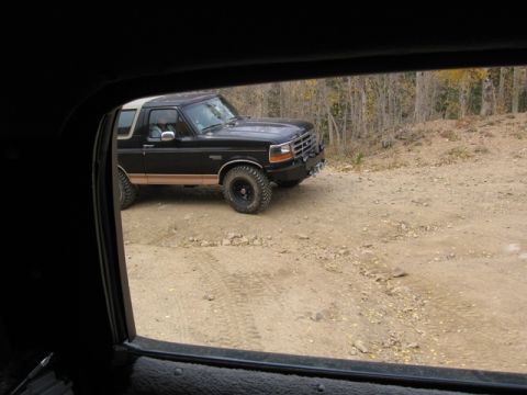 1996 Ford Bronco - A black Ford Bronco with a tan stripe is parked on a dirt road with trees in the background. The view is from inside another vehicle, looki…