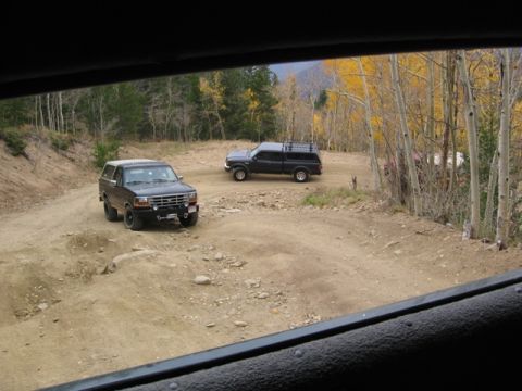 1996 Ford Bronco - Two dark-colored SUVs are parked on a dirt road in a wooded area with autumn foliage. The SUV in the foreground is a dark-colored Bronco, a…