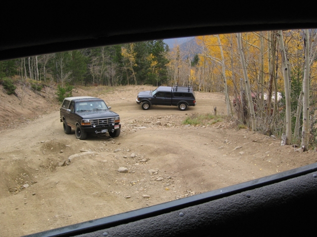 Two dark SUVs on a dirt road in a forest with fall colors.