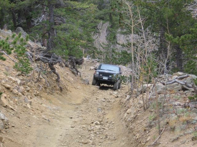 Black Jeep Cherokee driving up a steep, rocky off-road trail in a forest.