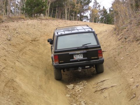 1996 Ford Bronco - A black Jeep Cherokee is driving up a steep, rutted dirt road with sparse trees on either side.