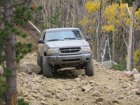 1996 Ford Bronco - A silver Ford Explorer is driving on a rocky, off-road trail surrounded by trees with yellow and green foliage. The vehicle is angled towar…