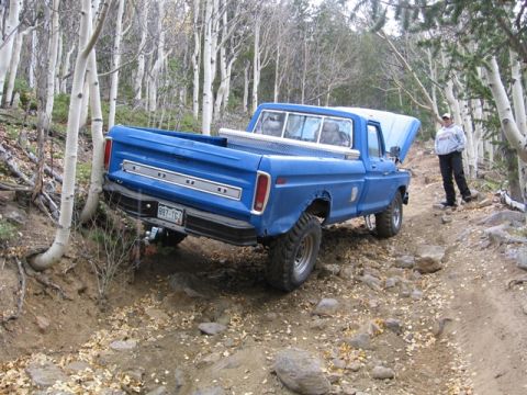 1996 Ford Bronco - A blue Ford pickup truck is shown from a rear three-quarter angle, tilted on a rocky, wooded trail. The hood is open, and a person stands t…