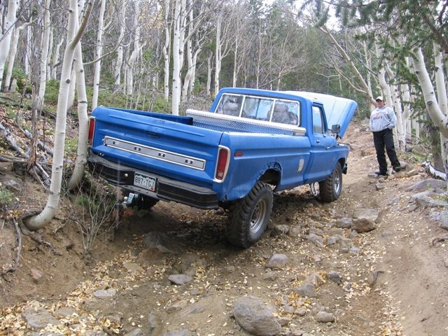 Blue Ford pickup truck tilted on a rocky trail with the hood open. A person stands nearby.