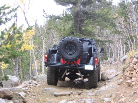 1996 Ford Bronco - The rear view of a modified black SUV is shown navigating a rocky, off-road trail in a wooded area with autumn foliage. A large spare tire…