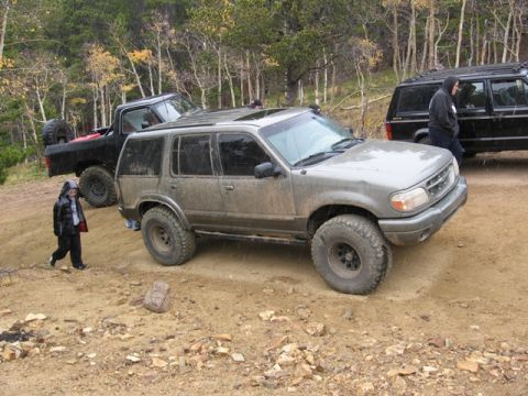 1996 Ford Bronco - A muddy, lifted Ford Explorer is parked on a dirt trail next to a black pickup truck. A child in a rain jacket walks in the foreground.