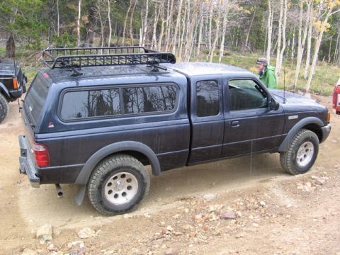 1996 Ford Bronco - A dark blue Ford Ranger pickup truck with a camper shell and roof rack is parked on a dirt road in a wooded area. The truck has off-road ti…