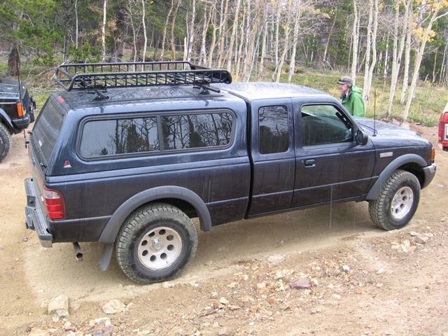 Dark blue Ford Ranger pickup truck with camper shell and roof rack on a dirt road.