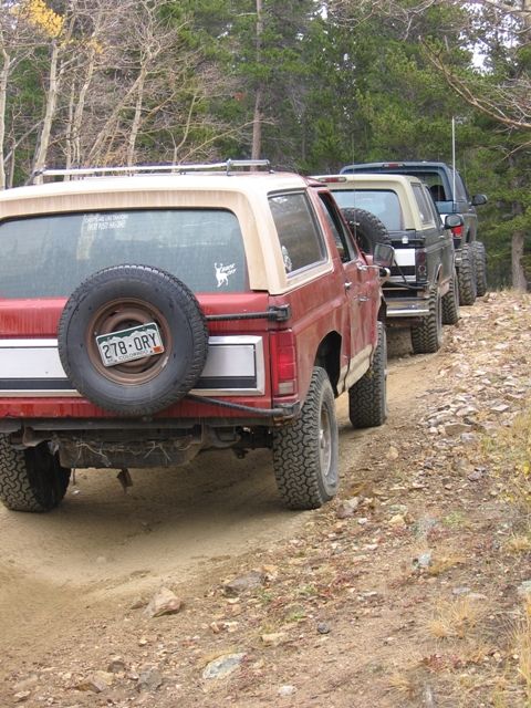 1996 Ford Bronco - A line of Ford Broncos are driving on a dirt road in a forest. The lead vehicle is a red Bronco with a tan top, showing its rear and spare…
