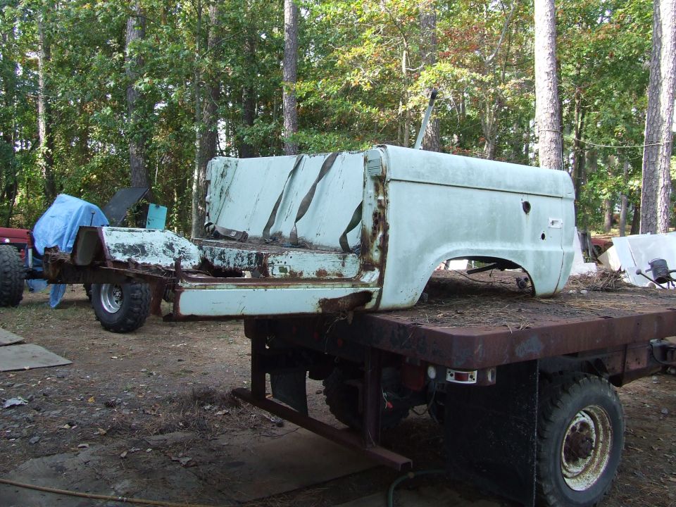 Weathered light blue Ford Bronco body on a rusty flatbed trailer in a wooded area.