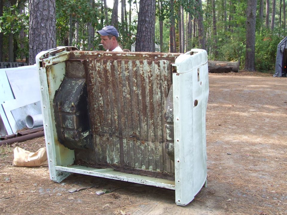 Rusted truck bed with a man smoking in the background, outdoors.